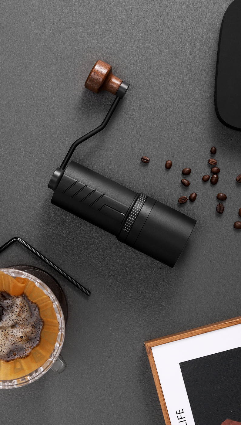 Top-down view of the VEVOK CHEF O20 Manual Coffee Grinder on a gray table, surrounded by freshly brewed pour-over coffee, scattered coffee beans, and part of a picture frame, showcasing its aesthetic appeal and coffee brewing excellence.