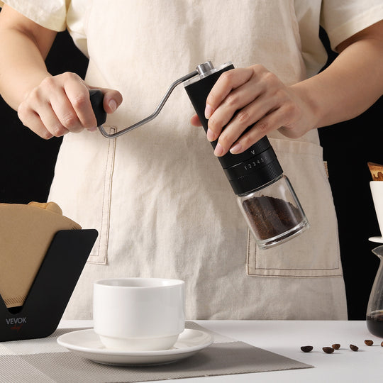 A barista grinding coffee beans with the VEVOK CHEF O6 Slim Manual Coffee Grinder Glass Jar, surrounded by a coffee cup and coffee filter holder, highlighting the grinder's glass jar option for brewing enthusiasts.