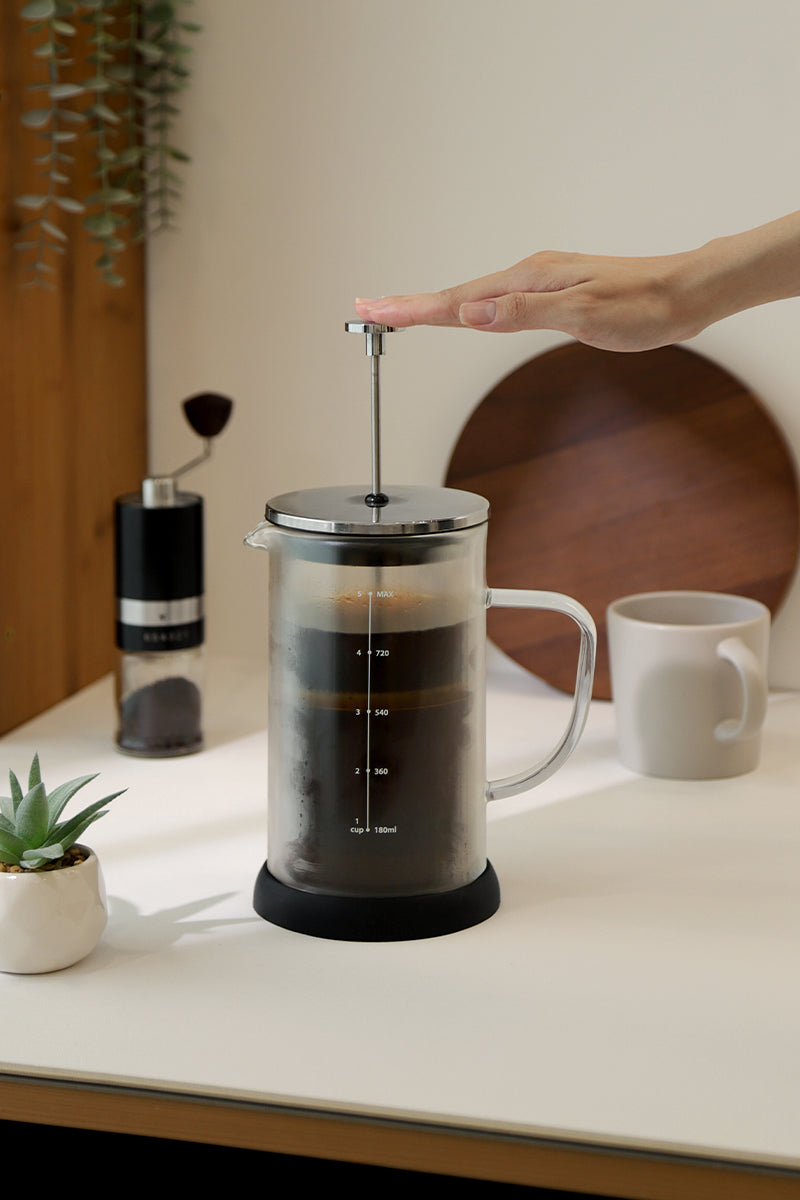 Brewing cold brew coffee with a French press, hand pressing down slowly; VEVOK CHEF O6 manual grinder and coffee cup on the table in a soft-toned setting.