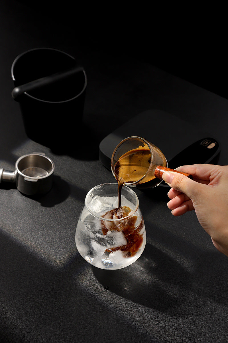 Pouring espresso into a glass with ice to make an iced Americano, with VEVOK CHEF espresso portafilter, knock box, and digital scale on a dark background.