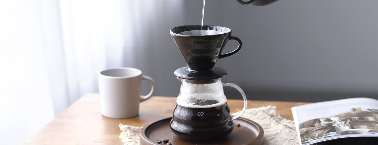 The NERA Ceramic V-Shaped Pour Over Coffee Dripper in use, placed on a glass coffee server with hot water being poured over the coffee grounds. The brewing process is captured with coffee liquid filling the glass server. On the table, a wooden tray, gray mug, and an open magazine add to the cozy, stylish atmosphere. VEVOK CHEF's NERA dripper ensures precision and flavor in every pour.