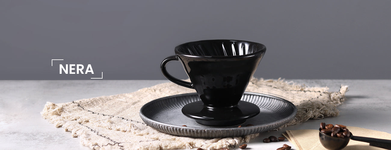 The NERA Ceramic V-Shaped Pour Over Coffee Dripper elegantly displayed on a gray tray with a beige tablecloth on a light gray table. The background features a dark gray wall, with coffee filters and beans placed beside the dripper. This scene highlights the minimalist beauty and functional design of the NERA dripper by VEVOK CHEF.