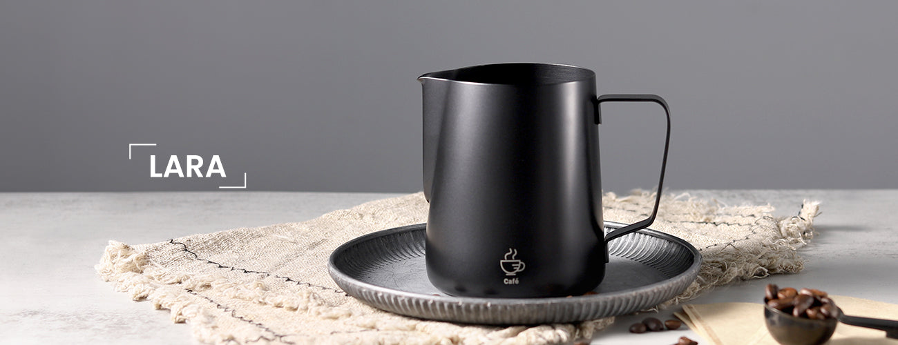 The VEVOK CHEF LARA Milk Frothing Pitcher displayed on a stone table with a gray wall backdrop. The scene includes a plate, napkin, coffee spoon, coffee beans, and filters, showcasing the pitcher’s sleek and stylish design as part of a refined coffee setup.