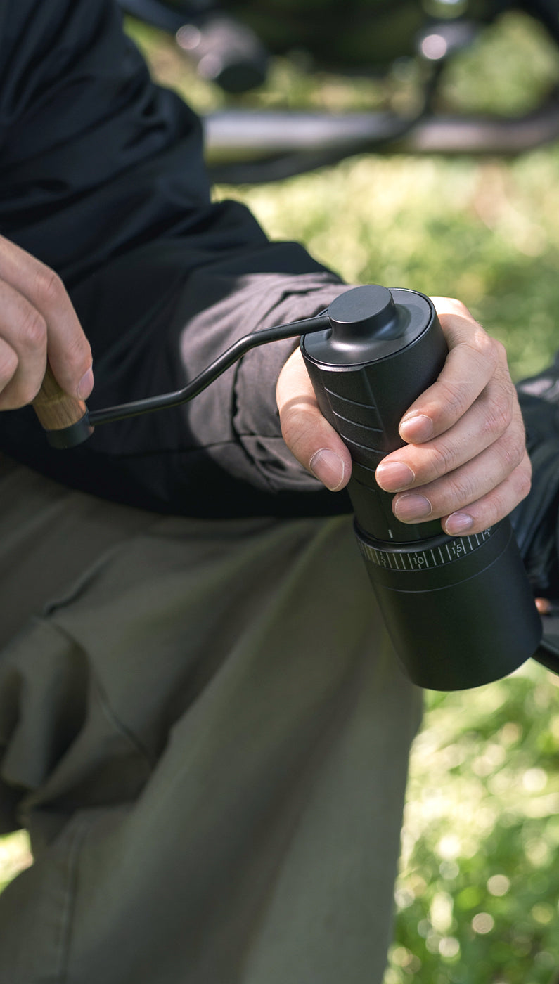 A person sitting on a camping chair with grass in the background, holding and using the VEVOK CHEF O20 Manual Coffee Grinder, showcasing its portability and suitability for outdoor adventures.