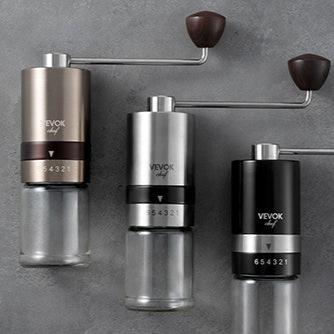 The VEVOK CHEF O6 Manual Coffee Grinder in gold, silver, and black placed on a gray tabletop, surrounded by coffee beans and brewing tools, highlighting its premium design and versatile color options.