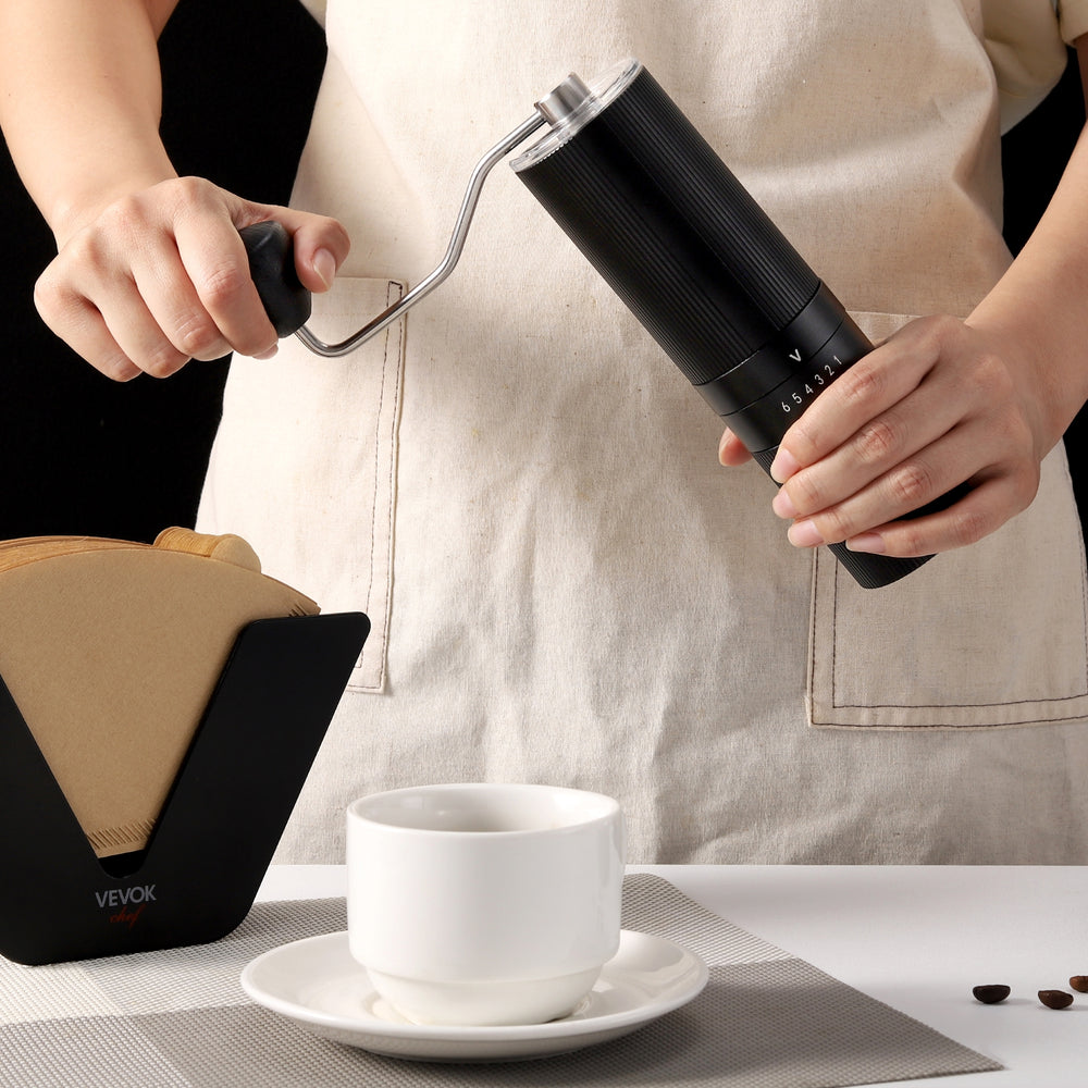 A barista grinding coffee beans with the VEVOK CHEF O6 Slim Manual Coffee Grinder, surrounded by a coffee cup and coffee filter holder, showcasing its professional-level performance.