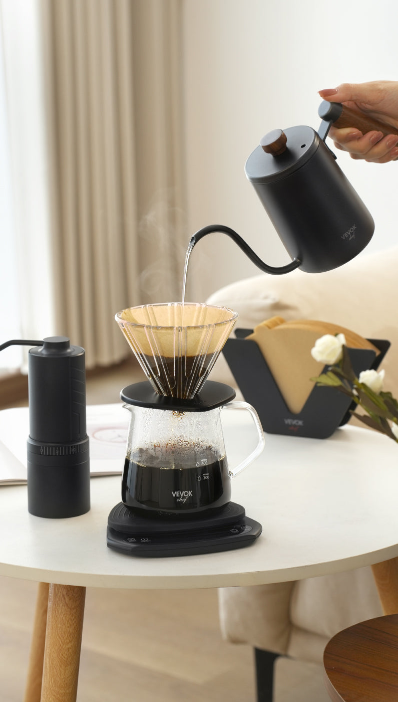 Elegant coffee setup featuring the VEVOK CHEF Pour Over Coffee Maker Set on a white round table in a cream-toned living room. A long-spout kettle pours water into the dripper, surrounded by coffee tools like the O20 grinder, filter holder, and fresh flowers. Ideal for specialty coffee lovers.