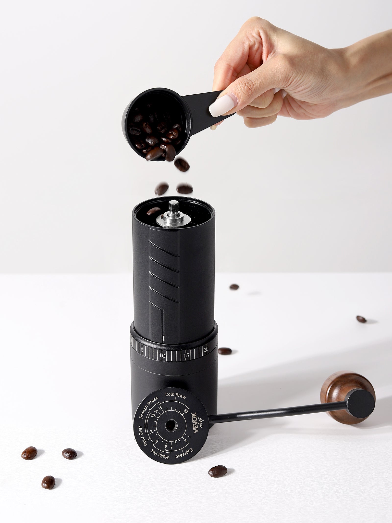 A hand opening the bean hopper of the VEVOK CHEF O20 manual coffee grinder while adding fresh coffee beans. The inside of the lid displays a grind size adjustment guide, ideal for espresso, pour-over, French press, and cold brew coffee lovers.